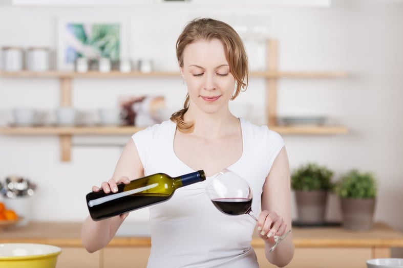 Attractive young woman standing pouring herself a glass of red wine in the kitchen with a smile of anticipation.jpeg